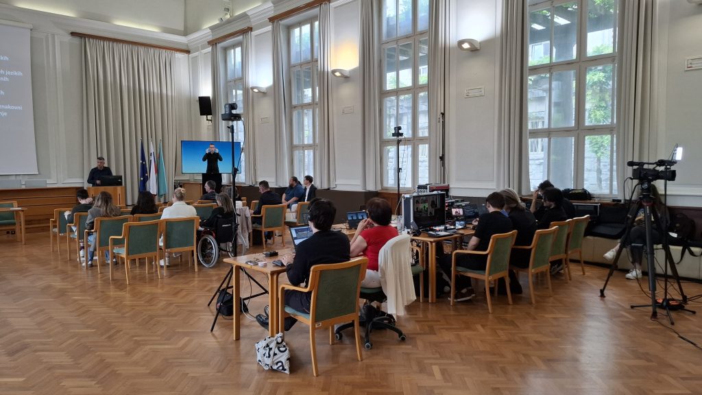A conference room filled with attendees seated at desks, working on laptops. Video cameras are positioned among the audience to record the event. In the back corner, a screen displays a sign language interpreter, ensuring accessibility for participants with hearing impairments.
