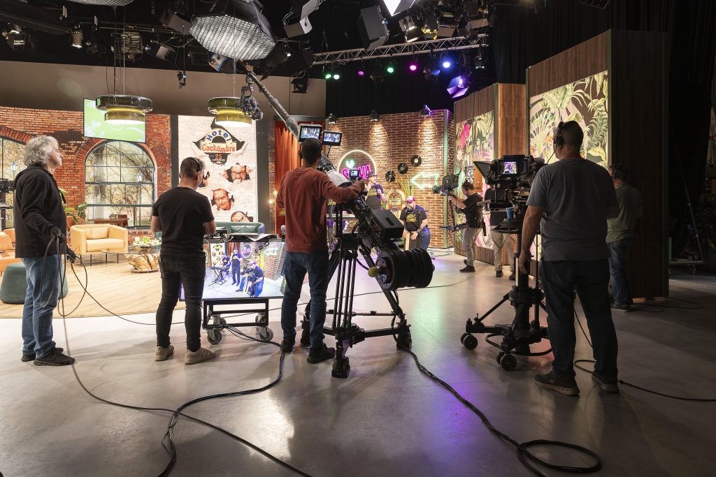 A behind-the-scenes view of a television studio during the filming of a show. Several camera operators and crew members are seen working with professional filming equipment, including cameras, monitors, and boom microphones. The set is decorated with vibrant lighting, brick walls, neon signs, and furniture arranged to resemble a cozy talk show or entertainment set. Some individuals are seated on stage while the crew captures the scene.