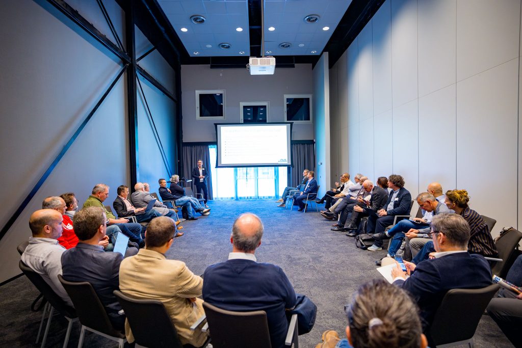 A conference room setup during an industry unconference session, where a presenter stands at the front beside a projection screen addressing a seated audience arranged in a semi-circle. Attendees, mostly professionals in business attire, are listening attentively, some taking notes or using laptops. The room features modern architecture with high ceilings, large wall panels, and soft lighting, creating a focused and collaborative discussion environment.