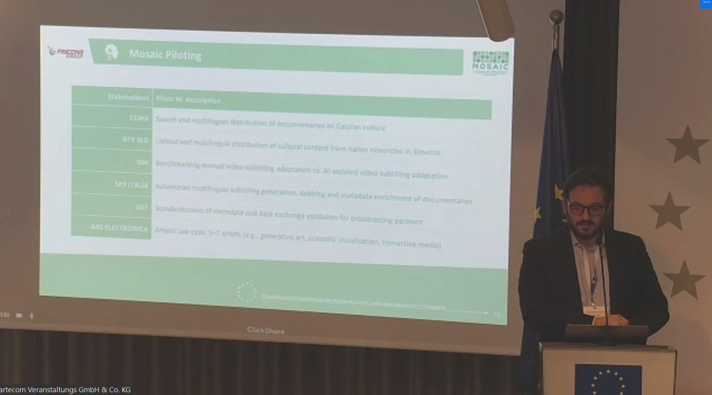 A man stands at a podium giving a presentation, with a large projected slide behind him titled “Mosaic Piloting.” The slide lists stakeholders and pilot use cases, while EU flags are visible to the right of the speaker.